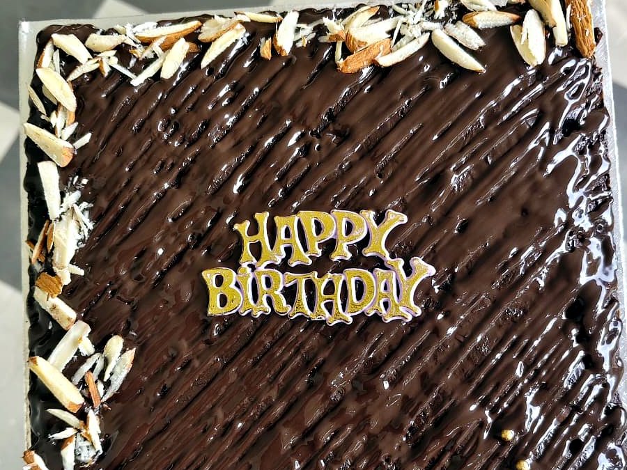 Square chocolate birthday cake with glossy ganache, almond flakes and a gold Happy Birthday topper