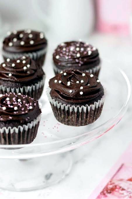 Chocolate cupcakes with swirled dark frosting and white pearl sprinkles on a glass stand