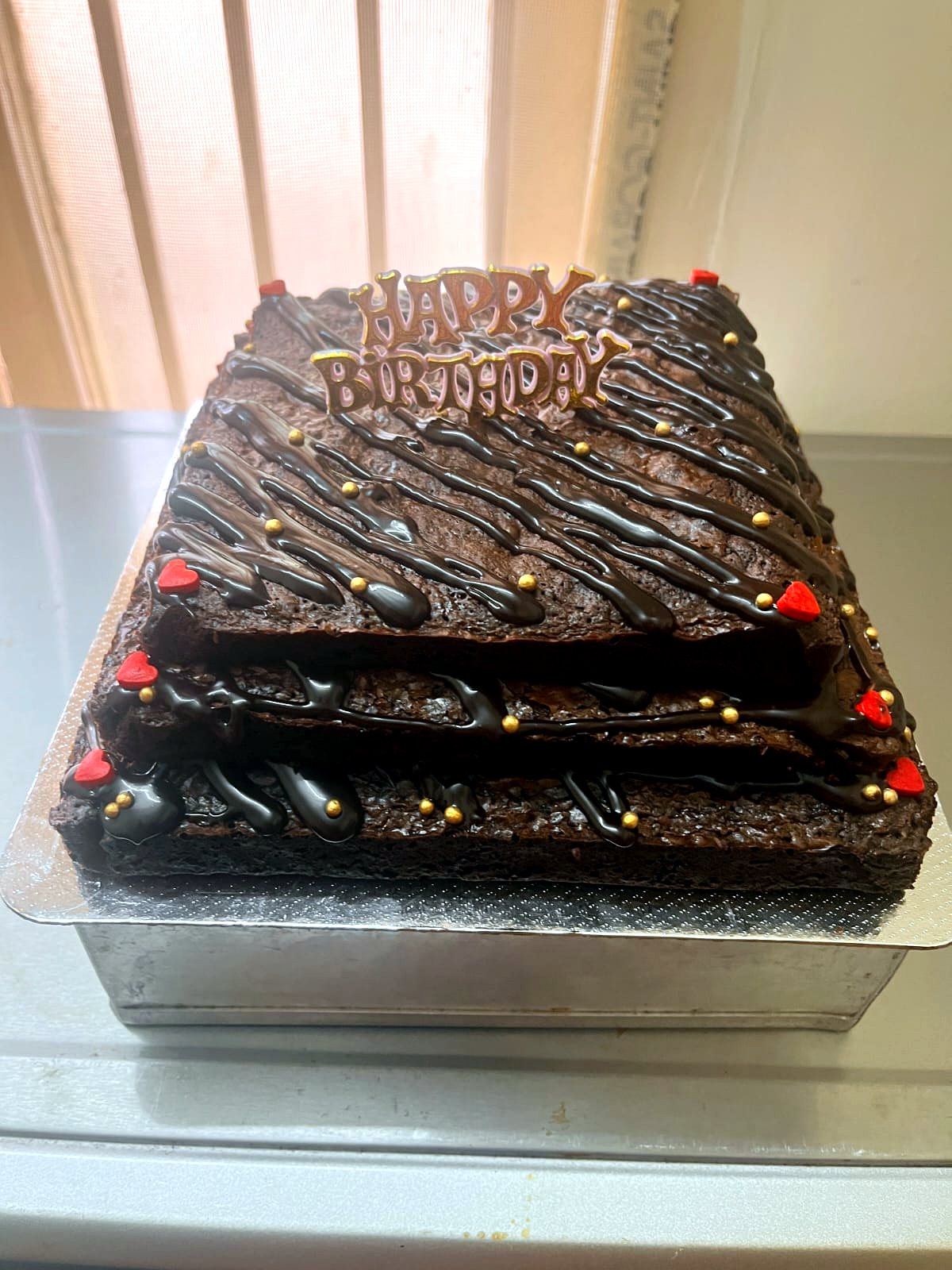 Three-tier birthday cake made of stacked brownie layers with chocolate drizzle, gold pearls and red hearts